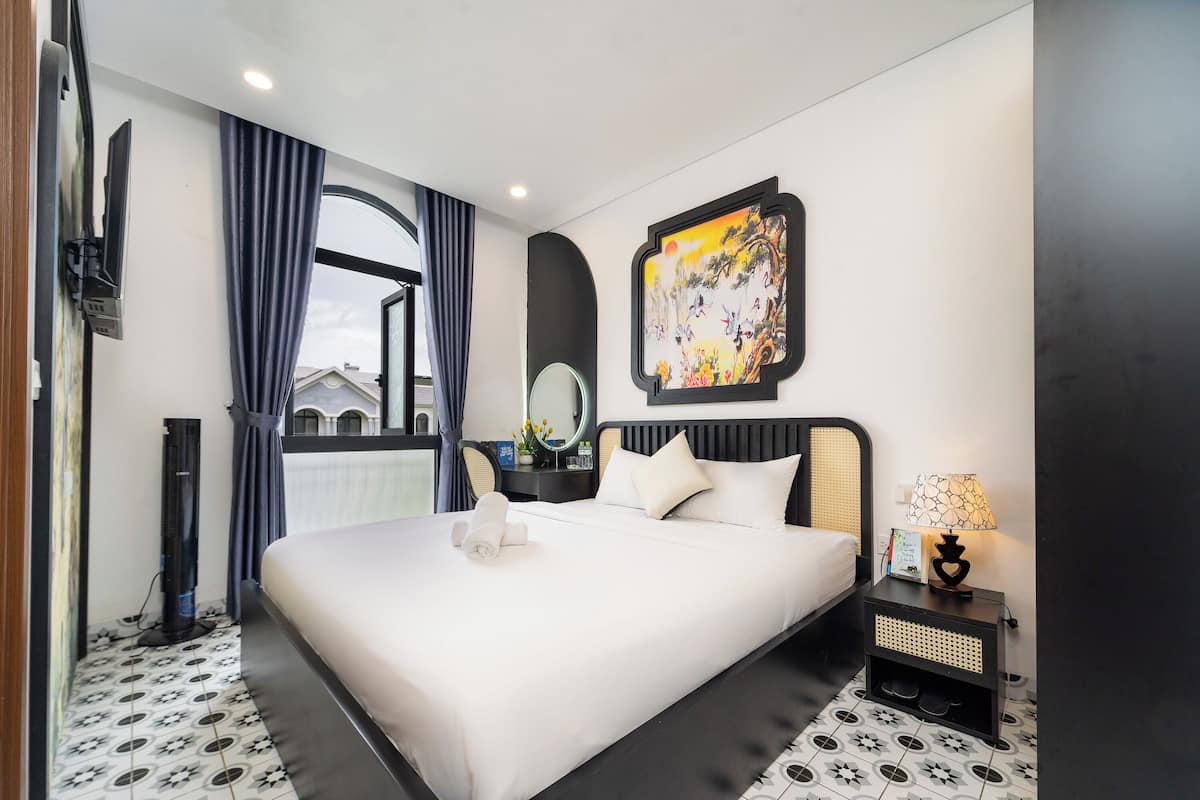 A hotel room with a large bed, a bedside table with a lamp, a wall-mounted TV, and a window with curtains.