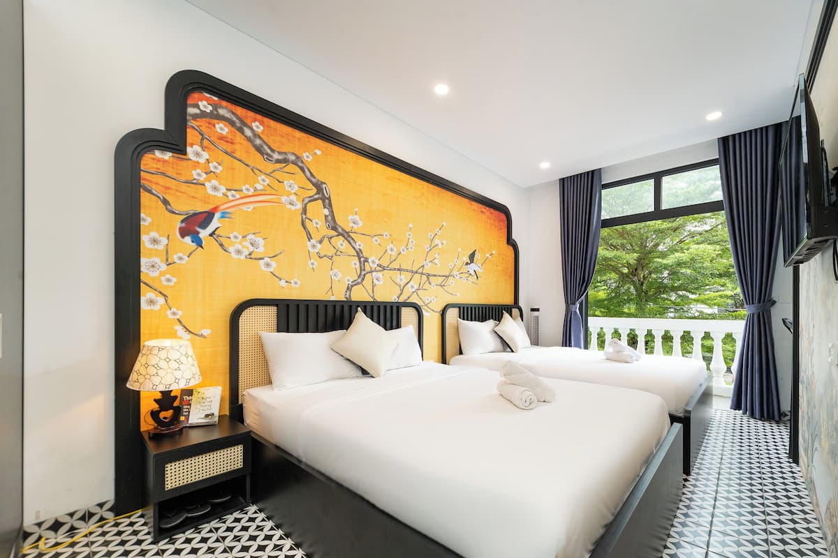 A hotel room with two beds, a decorative wall mural, a nightstand with a lamp, and a view of greenery outside.