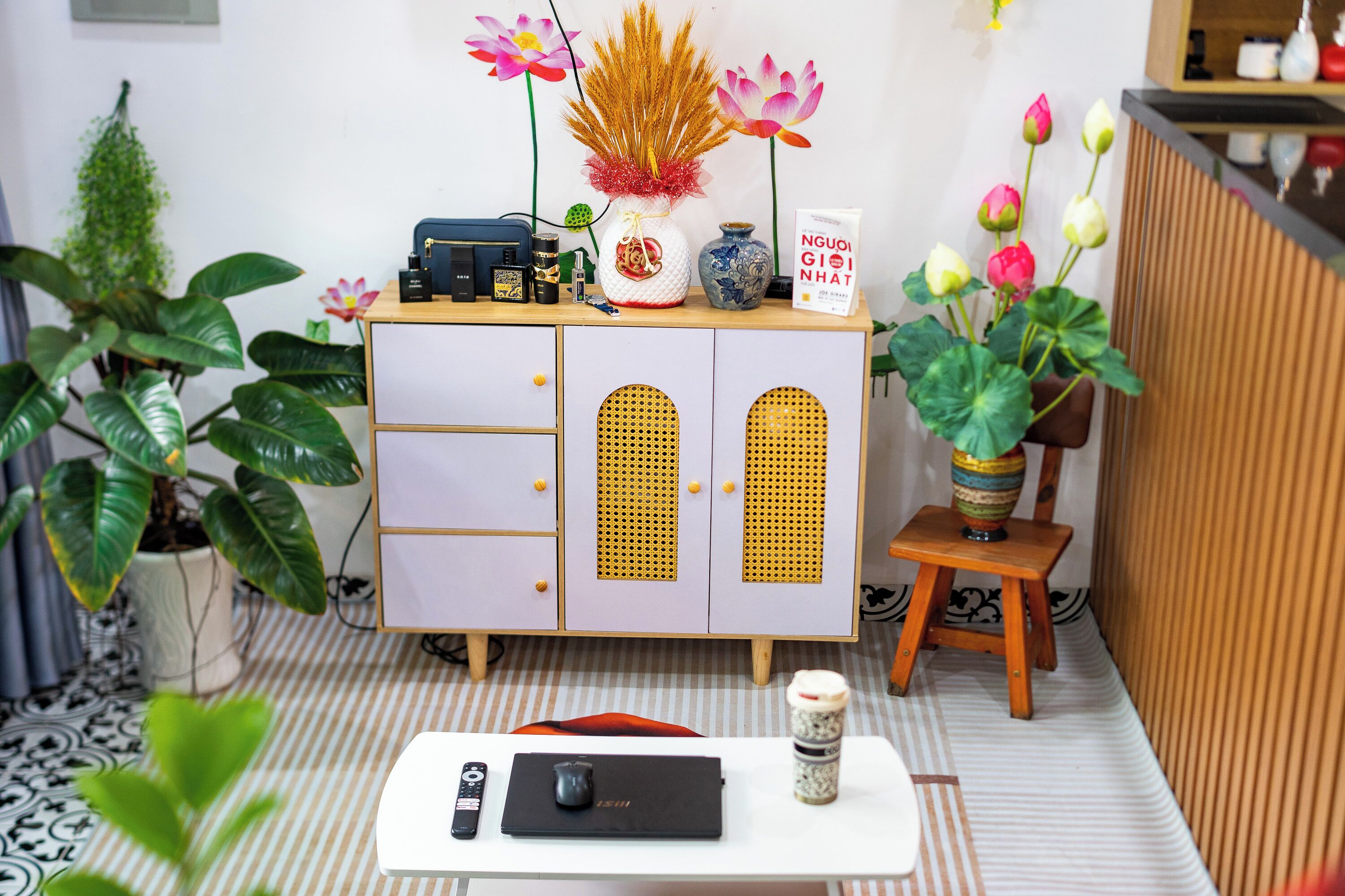 Una habitación ordenada con una mesa blanca, un mueble con cajones, un taburete pequeño y plantas en macetas.