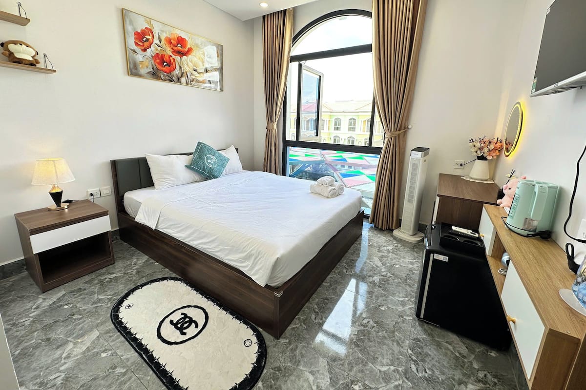 A hotel room with a bed, bedside lamp, a small table, a desk with a coffee machine, a mirror, and a window with curtains.