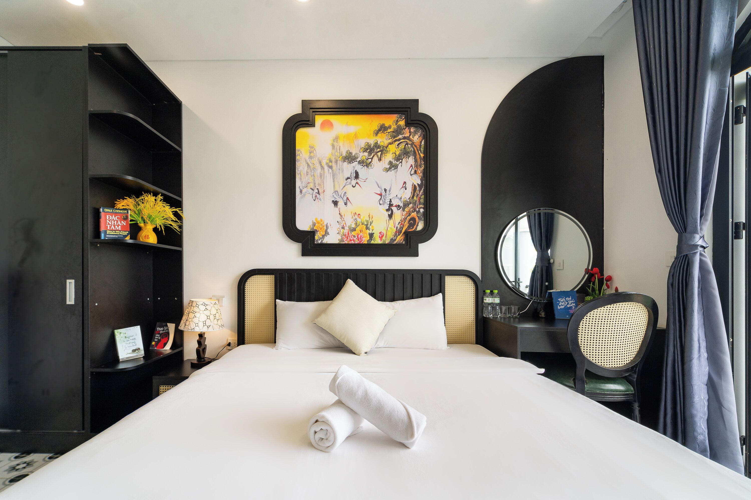 Habitación de hotel con una cama bien hecha, una mesita de noche con una lámpara, una estantería, un escritorio con un espejo y un cuadro en la pared.