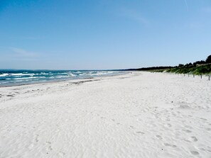 Uma extensa praia de areia com ondas suaves, céu claro e uma faixa de vegetação à direita.
