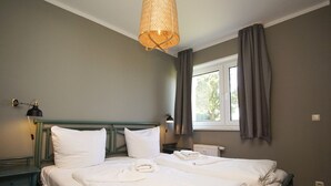A double bed with white linens and towels, a green headboard, a bedside lamp, and a window with curtains.
