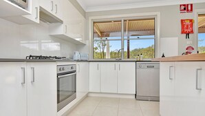 Une cuisine moderne avec des meubles blancs, une cuisinière à gaz, un four, un lave-vaisselle et une fenêtre donnant sur les arbres.