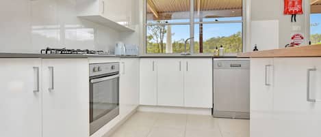 A modern kitchen with white cabinets, a gas stove, an oven, a dishwasher, and a window with a view of trees.