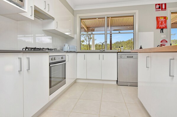 A modern kitchen with white cabinets, a gas stove, an oven, a dishwasher, and a window with a view of trees.