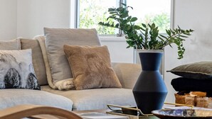 A living room with a sofa, cushions, a vase with a plant, a wooden table with books and a bowl, and a window with a view of greenery.