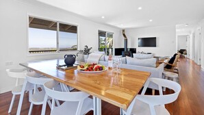 A modern living room with a wooden dining table, white chairs, a sofa, and a TV mounted on the wall.