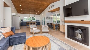 A spacious living room with a large TV, a fireplace, and a dining area with a kitchen in the background. - Beautiful Riverfront Retreats | Sleeps 24 w/ Hot Tubs, Firepits & Scenic Views (Shenandoah)