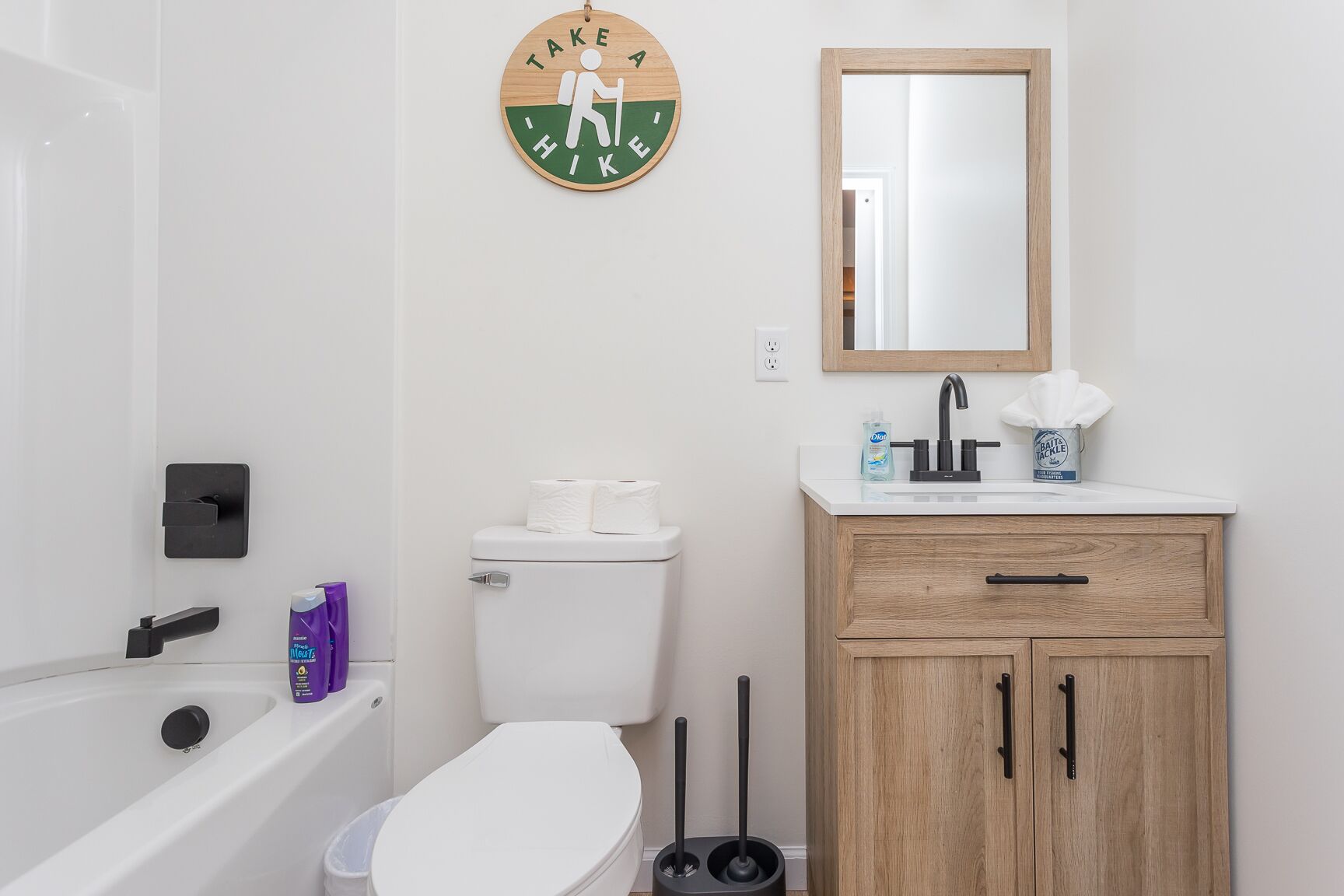 A bathroom with a toilet, sink, and bathtub. There is a wooden cabinet with a mirror above the sink. A sign on the wall reads "TAKE A HIKE".