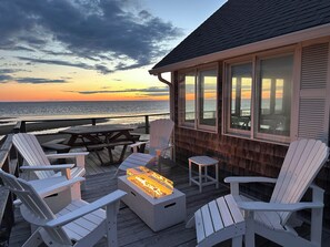 Eine Holzterrasse mit Adirondack-Stühlen, einer Feuerstelle und Blick auf den Ozean bei Sonnenuntergang.