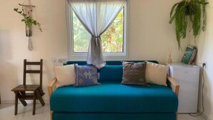 Un canapé de couleur turquoise agrémenté de coussins à motifs, une petite chaise en bois, un réfrigérateur blanc et une fenêtre ornée de rideaux légers.