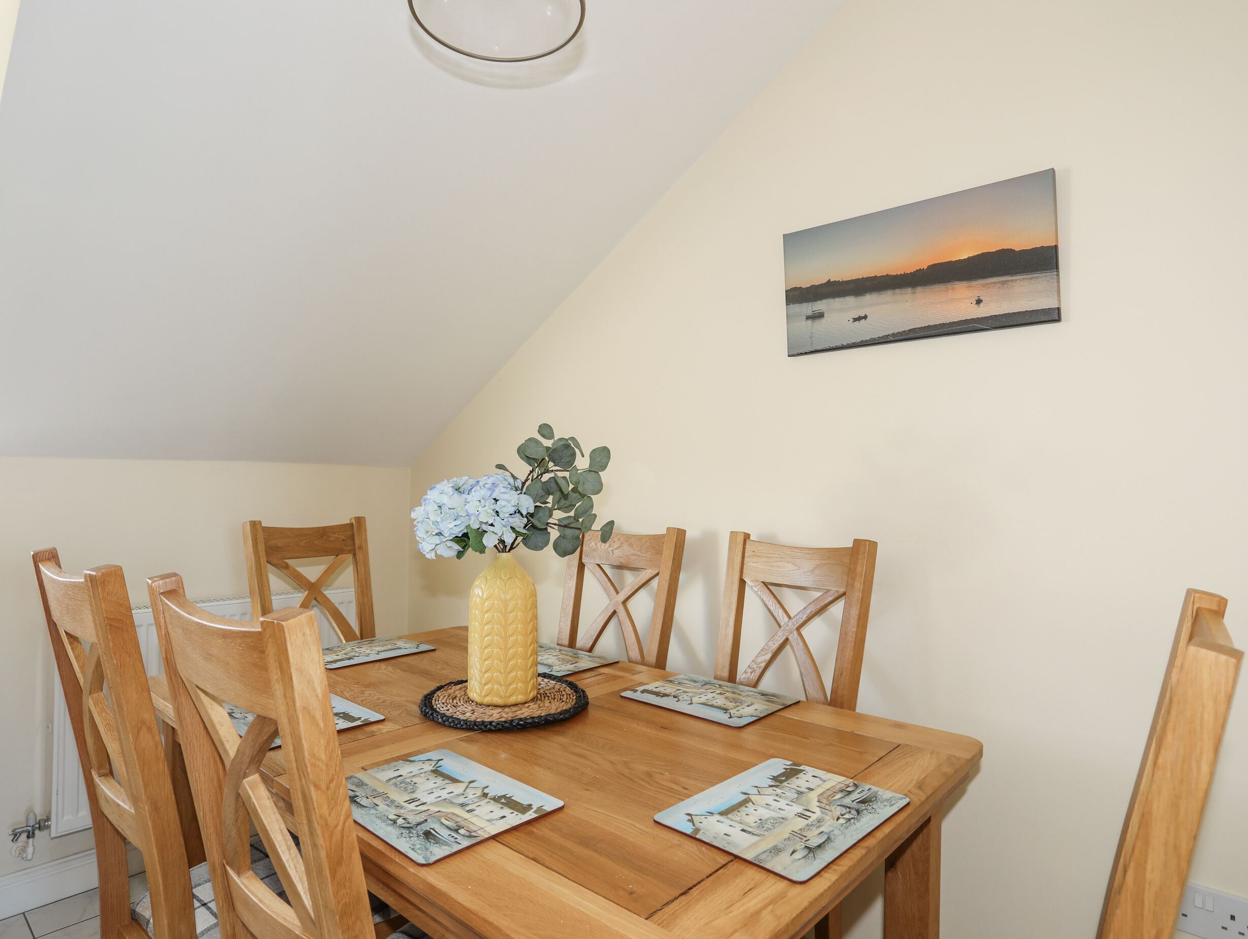 A dining table with wooden chairs, a vase with flowers, and a coastal sunset painting on the wall.