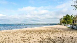 Une plage avec un rivage sablonneux, une étendue d’eau et une rangée de bâtiments bordée d’une promenade arborée.