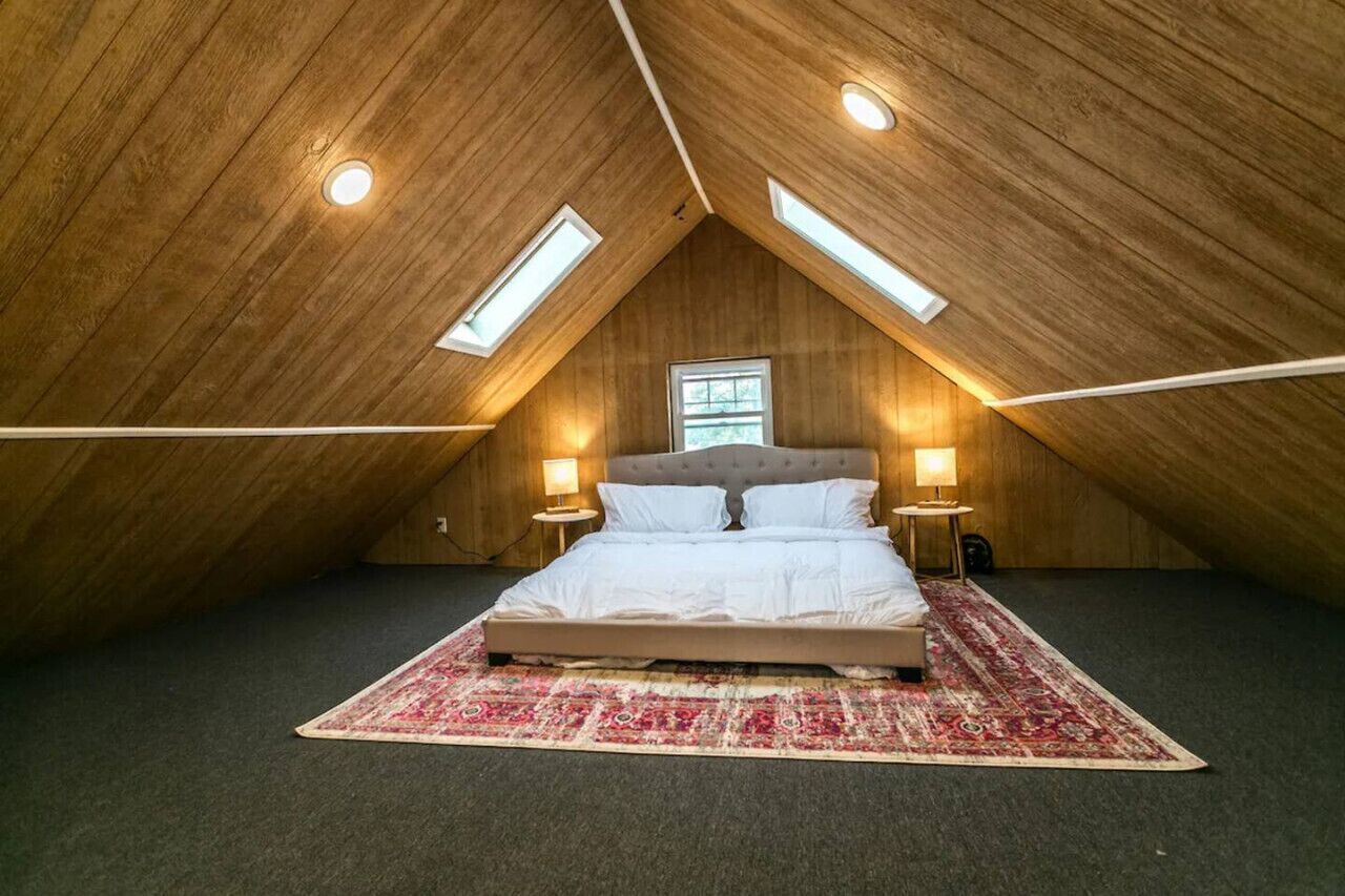 A bedroom with a sloped ceiling, a bed, two bedside lamps, a window, and a patterned rug.