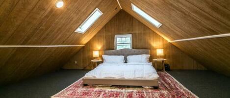A bedroom with a sloped ceiling, a bed, two bedside lamps, a window, and a patterned rug.