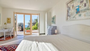 Un dormitorio con una cama grande, un escritorio y una silla. Se ve el exterior a través de una puerta de vidrio.