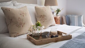A neatly made bed with floral pillows, a bedside table with a lamp, and a tray with a pitcher, mug, and fruit. - Butterwell Cottage at Collihole (Newton Abbot)