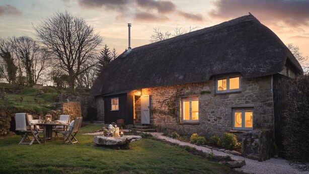 A stone cottage with a thatched roof, a stone chimney, and a well-lit outdoor seating area with a table and chairs. - Butterwell Cottage at Collihole (Newton Abbot)