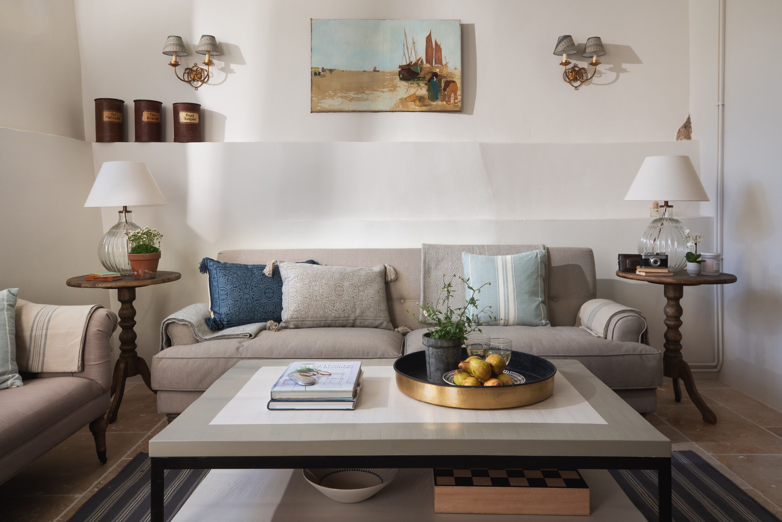 Un salon avec un canapé beige, deux tables d’appoint avec des lampes, une table basse avec un livre et un bol de fruits, et un tableau accroché au mur.