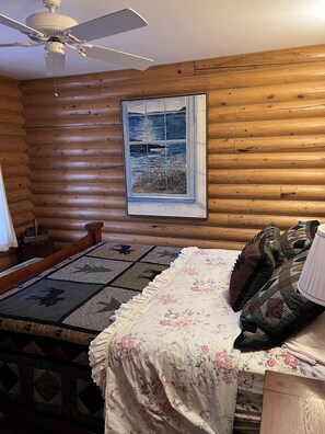 Une chambre dans une cabane en rondins, avec un lit, un ventilateur de plafond, une fenêtre ornée d’un tableau et une tête de lit en bois.