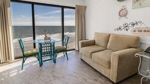 A room with a large window offering a view of the ocean, a beige sofa, a glass table with chairs, and a decorative wall piece.