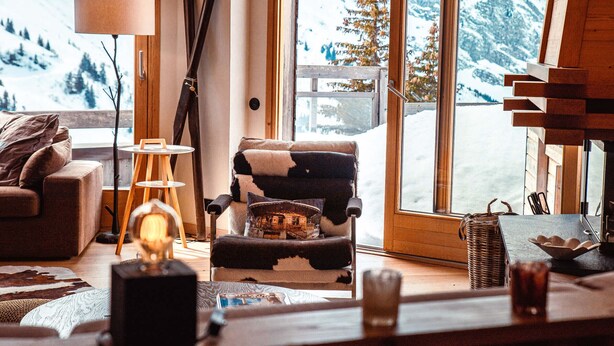 Ein gemütliches Wohnzimmer mit großem Fenster und Blick auf verschneite Berge, einem Kamin und einem Skiständer.