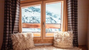 Ein gemütliches Zimmer mit großem Fenster und Blick auf einen verschneiten Berg, umrahmt von Karomustert-Vorhängen und zwei flauschigen Kissen.