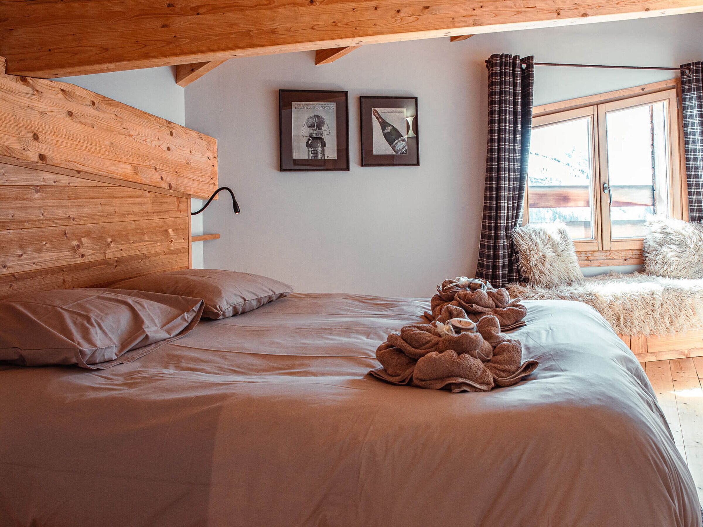 Un dormitorio con cabecera de madera, cama con ropa de cama marrón, ventana con cortinas de cuadros y dos cuadros enmarcados en la pared.