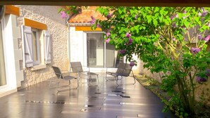 Un patio cubierto con pared de piedra, dos ventanas con contraventanas y un conjunto de muebles con sillas y una mesa.