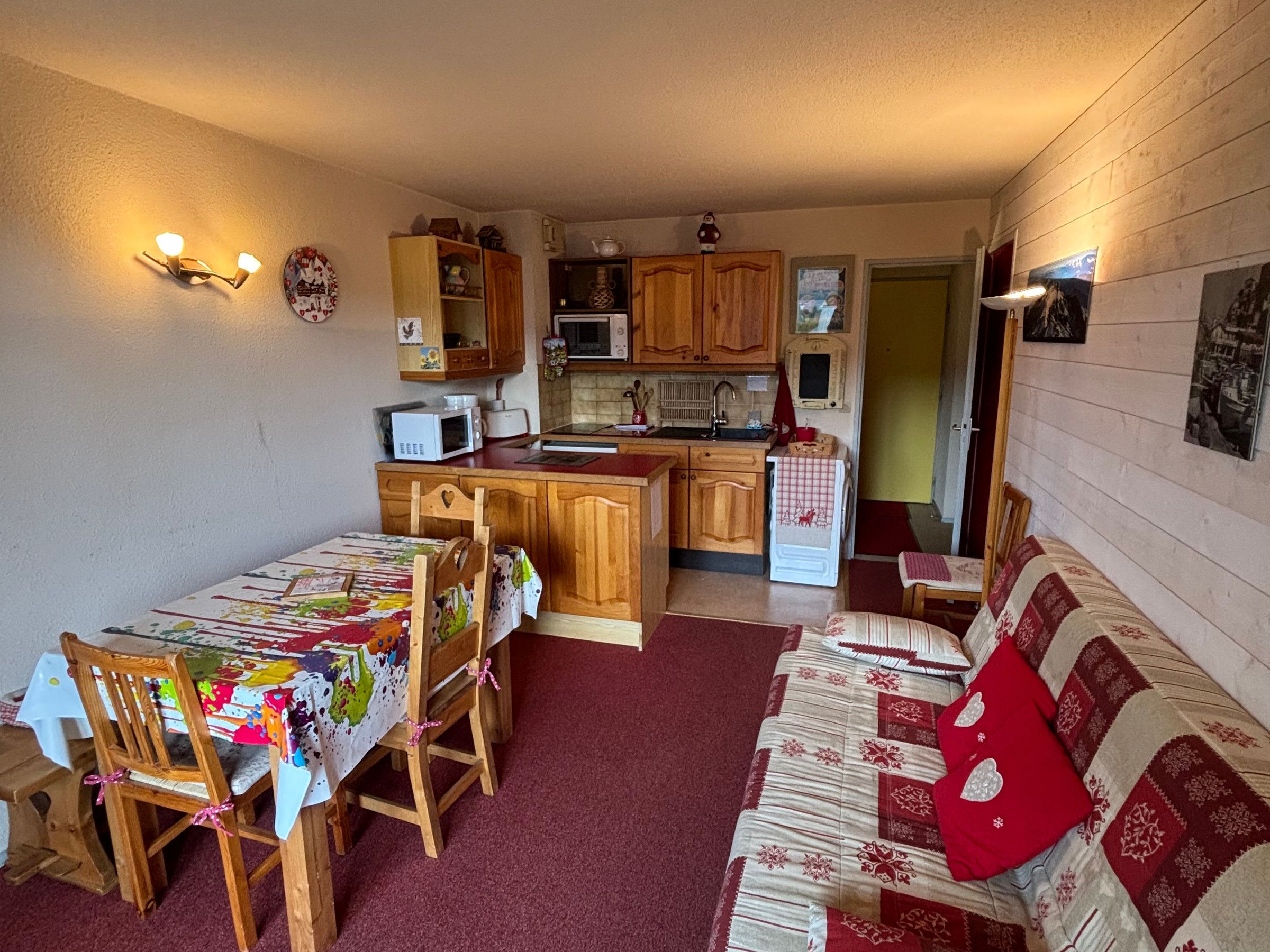 A compact kitchen with wooden cabinets, a microwave, and a sink. There is a dining area with a table and chairs, and a sofa with a red cushion.