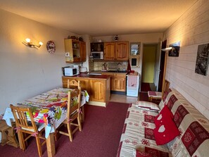 Une cuisine compacte avec des meubles en bois, un four  à micro-ondes et un évier. On y trouve un coin repas avec une table et des chaises, ainsi qu’un canapé agrémenté d’un coussin rouge.