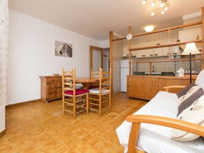 Ett kompakt rum med ett matbord och stolar i trä, en säng med kuddar, en köksdel med skåp och en väggklocka.