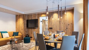 A modern living room with a wooden wall, a flat-screen TV, a dining area with a table set for four, and a sofa with cushions.