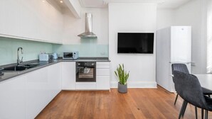 A modern kitchen with white cabinets, a built-in oven, a microwave, and a stainless steel range hood. There is a flat-screen TV mounted on the wall, a refrigerator, and a dining area with a table and chairs. - Luxury Apartment London Eye Westminister (London)
