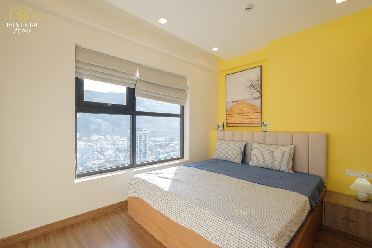A bedroom with a large window, a bed, a nightstand, and a wall-mounted lamp.