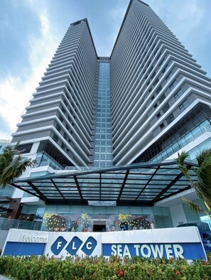 A modern high-rise building with a distinctive angular design, featuring a prominent entrance with a glass canopy and a sign reading "FLG SEA TOWER". - FLC Sea Tower - Quynhon Homes (Quy Nhon Nam)