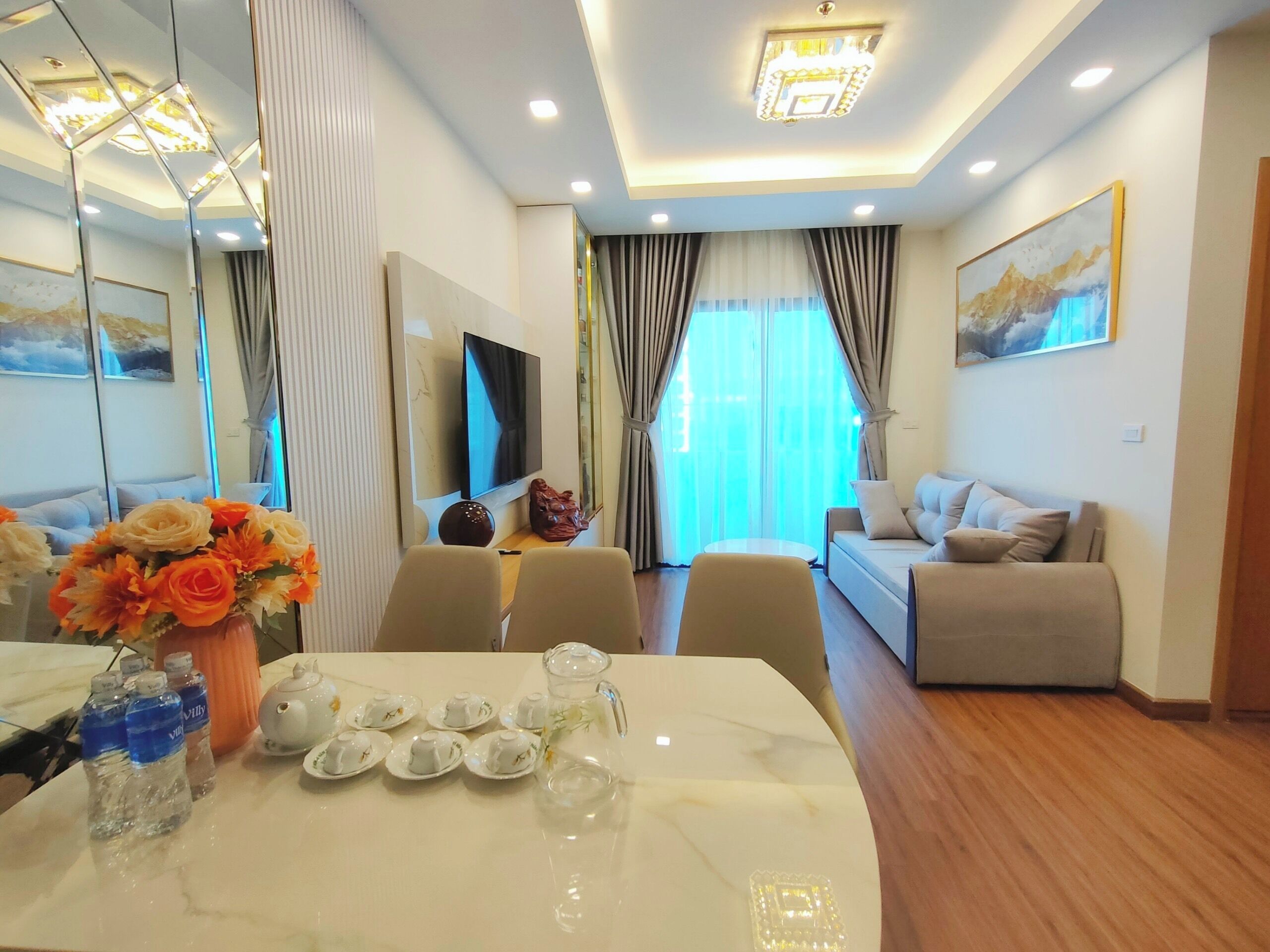 A modern living room with a sofa, a dining table with water bottles and teapots, and a TV.