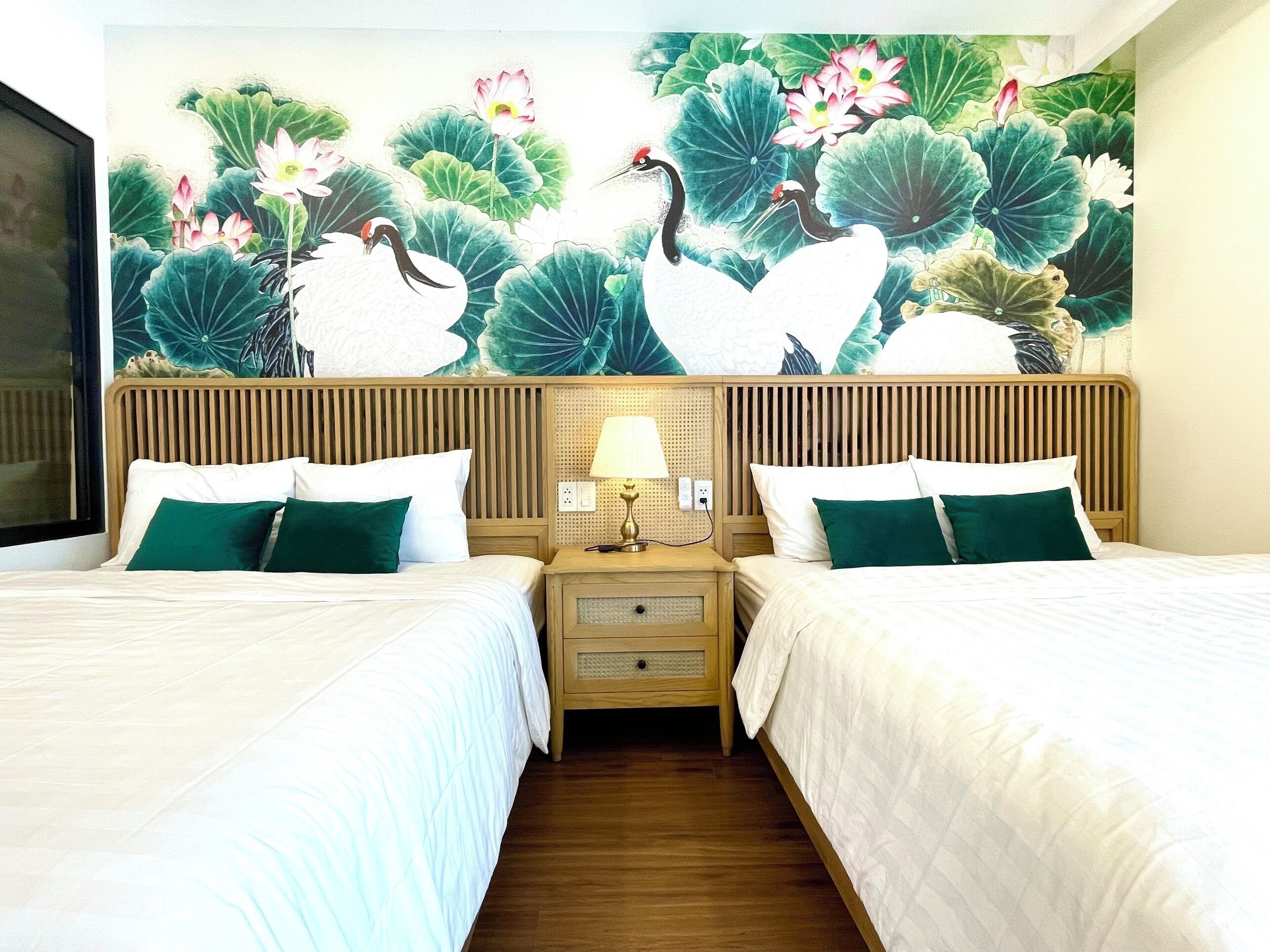 Une chambre d’hôtel avec deux lits, une tête de lit en bois, une table de chevet avec une lampe et une fresque murale représentant des fleurs de lotus et des oiseaux.