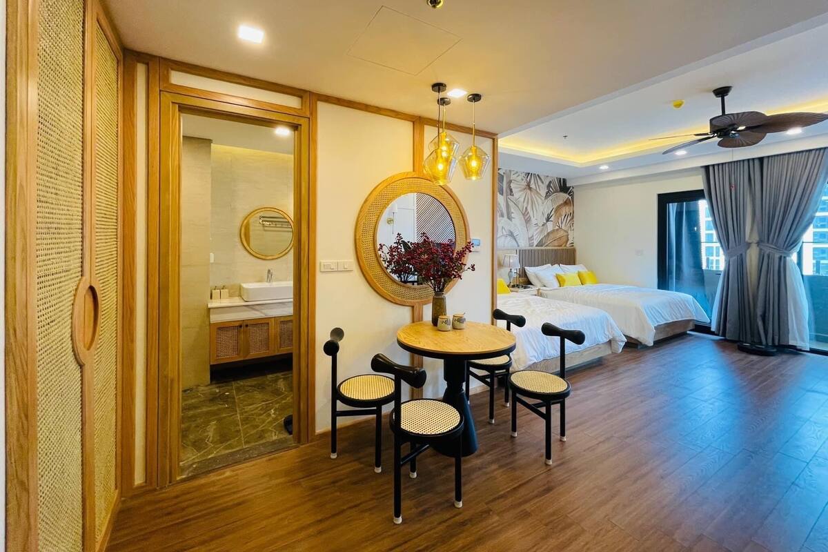 A hotel room with a wooden floor, a round dining table with chairs, a bed with white bedding, a ceiling fan, and a large window with curtains.