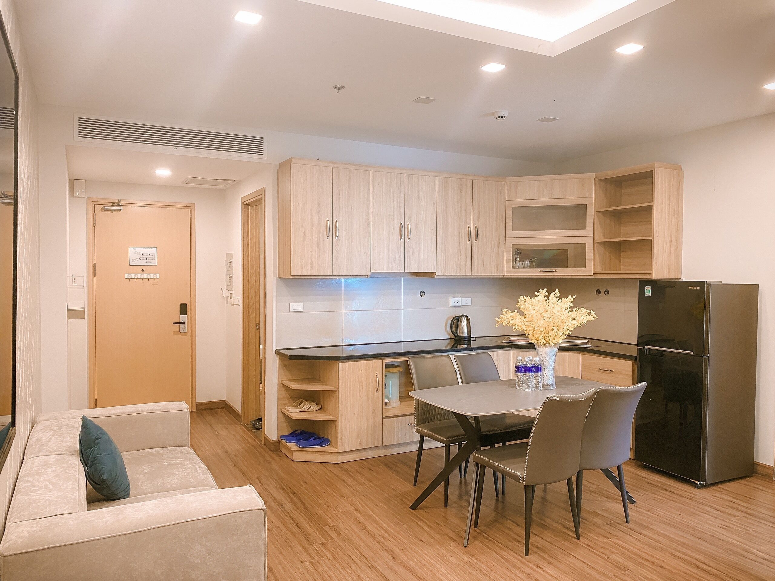 Cocina compacta con zona de comedor, armarios de madera, refrigerador negro y un florero con flores en la mesa.