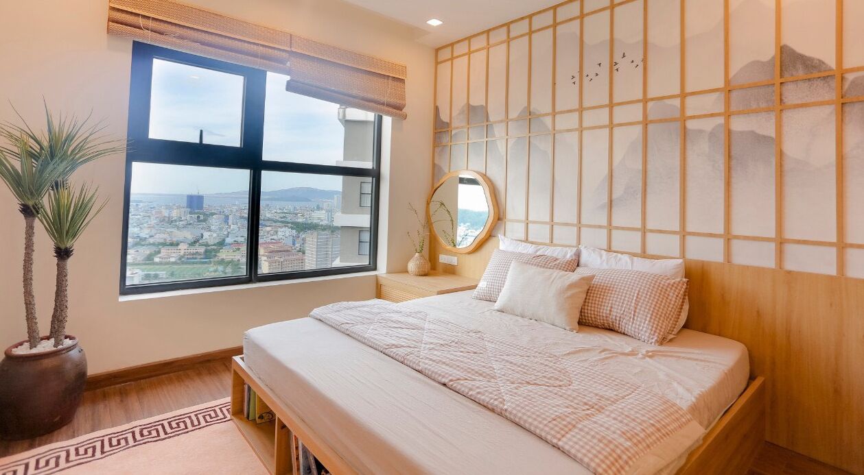 A bedroom with a large window, a bed, a bedside table, a mirror, and a decorative wall panel.