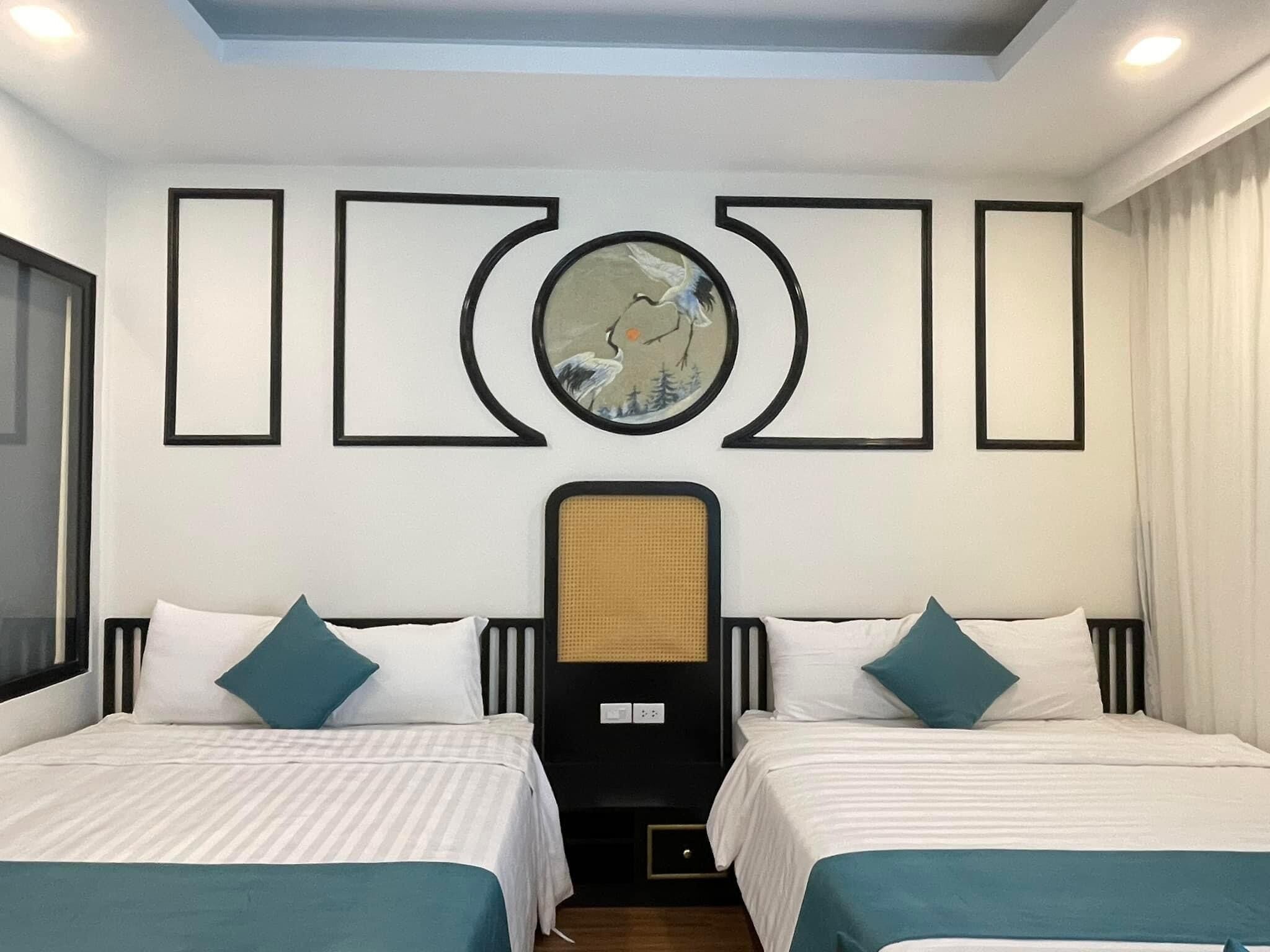 A hotel room with two beds, each with a headboard, white bedding, and teal pillows. There is a large circular artwork above the beds and a black panel with a woven texture in the center.
