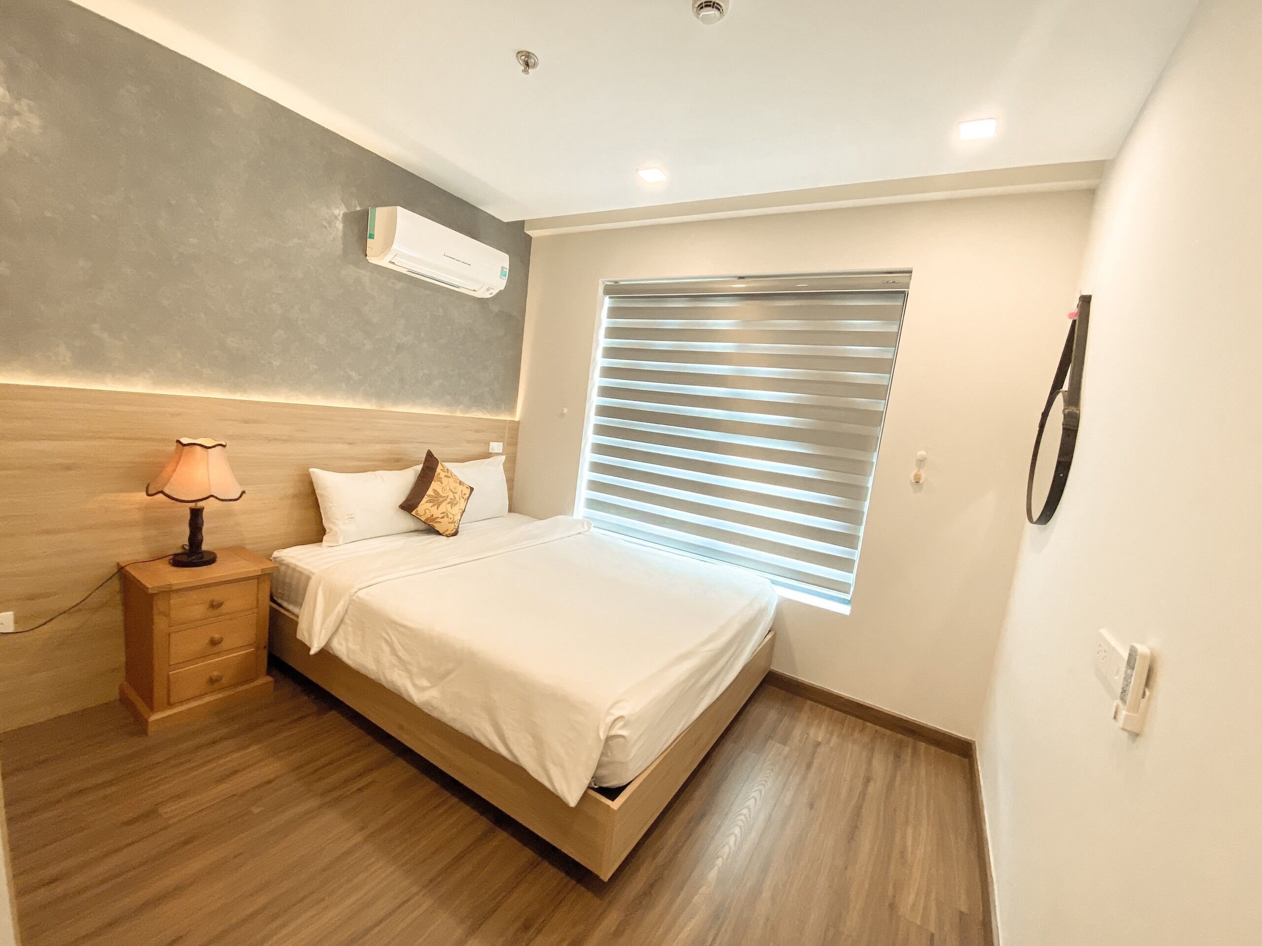 Ein Schlafzimmer mit Bett, Nachttisch, Lampe, Klimaanlage und einem Fenster mit Jalousien.