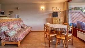 A cozy living room with a patterned sofa, a wooden dining table with chairs, a TV on a cabinet, and a large window with a view. - 6-Person Apartment at Foot of Slopes, Balcony, Equipped Kitchen (Morillon 1100)