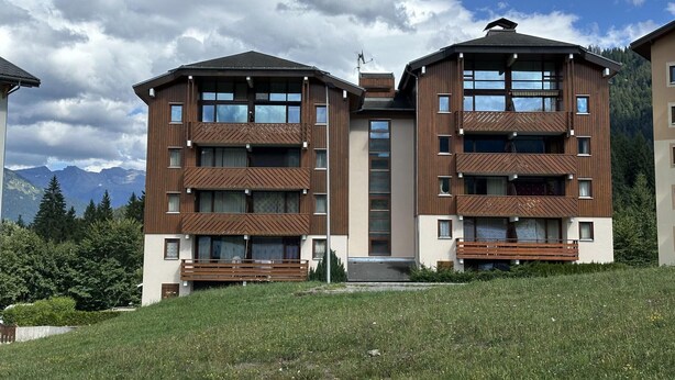 A modern multi-story building with balconies, large windows, and a view of mountains in the background. - 6-Person Apartment at Foot of Slopes, Balcony, Equipped Kitchen (Morillon 1100)