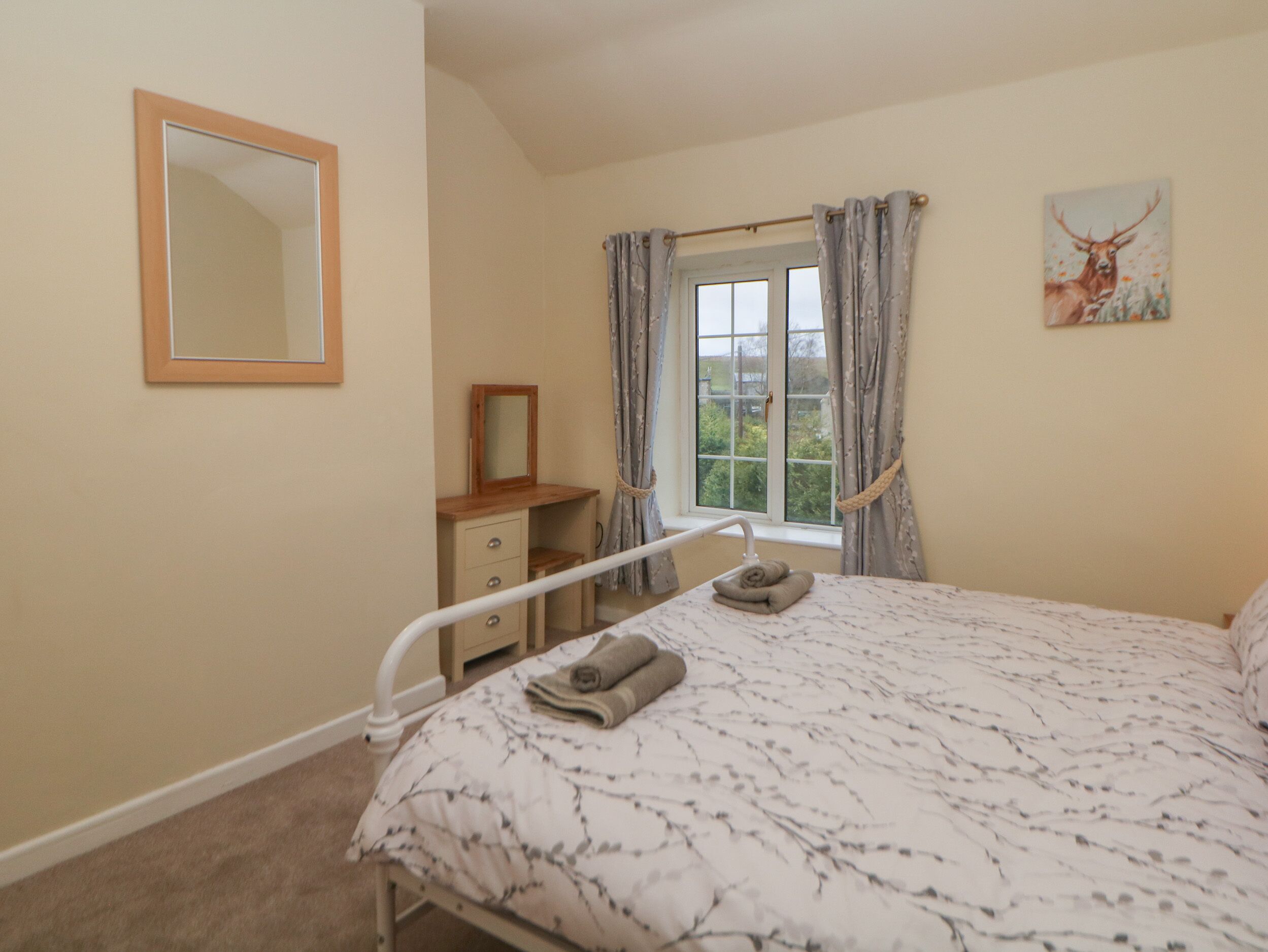 A bedroom with a bed, a bedside table, a mirror, a window with curtains, and a painting on the wall.