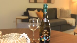 A bottle of Prosecco wine, two empty wine glasses, and a woven basket on a table in a living room with a couch and a painting in the background. - Palmira 1Bed/1Bath at the Tennis Village, Palmas del Mar (Humacao)