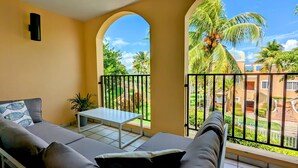 A balcony with a sofa, a small table, and a view of a landscaped garden with palm trees. - Palmira 1Bed/1Bath at the Tennis Village, Palmas del Mar (Humacao)