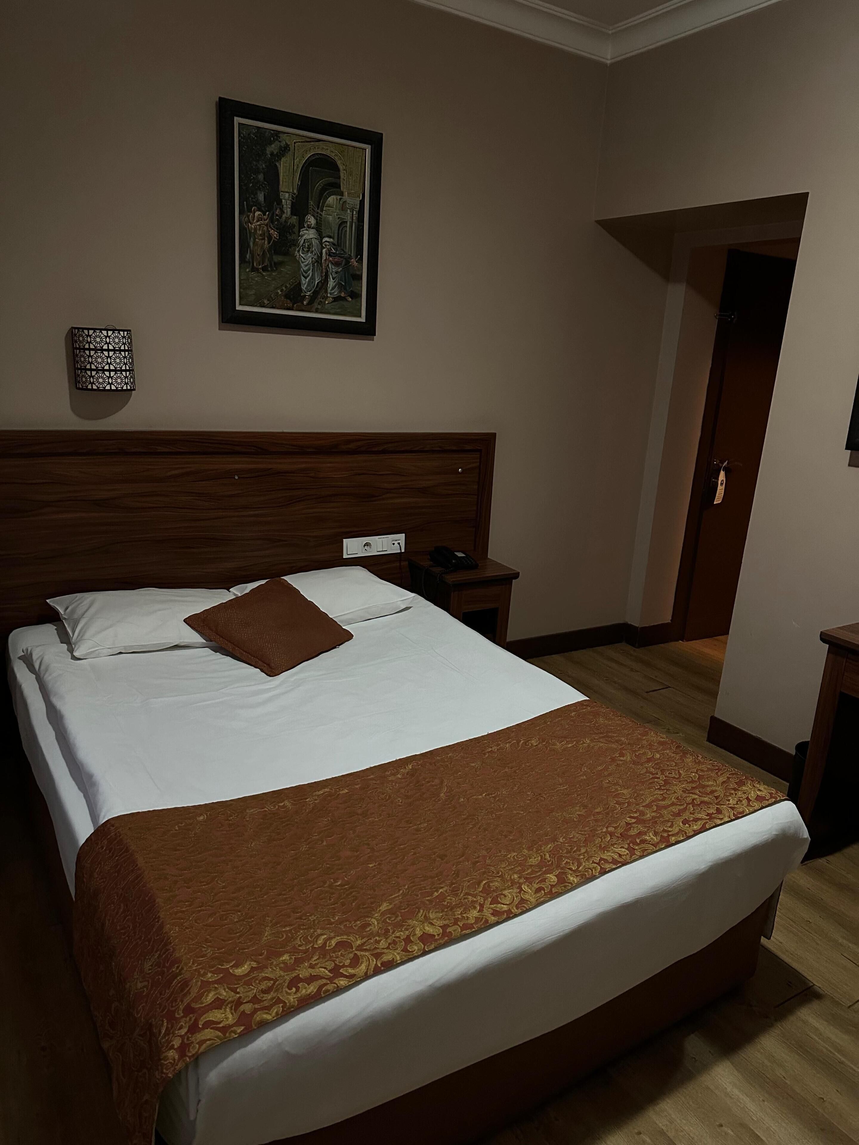 A neatly made bed with a brown blanket, a wooden headboard, a nightstand with a phone, and a framed picture on the wall.